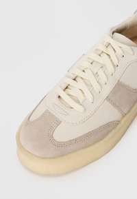 Beige suede sneaker with cream leather accents, round toe, flat laces, and a textured gum sole. Features contrast stitching details.