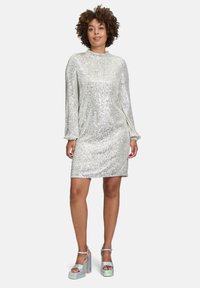 Srebrna sukienka z cekinami, z długimi rękawami i wysoką szyją. Zestawiona z masywnymi, iryzującymi sandałami na platformie. Gładka, odbijająca światło faktura.