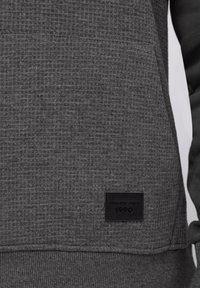 Prim-plan al unui hanorac din țesătură texturată gri închis, cu buzunar frontal și o etichetă neagră cu inscripția „ATHLETIC DEPT. 1990” pe marginea inferioară.