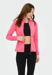 Giacca con zip color rosa acceso, collo alto, maniche lunghe e polsini elasticizzati. Indossata sopra una maglia nera. Tessuto liscio e leggero.