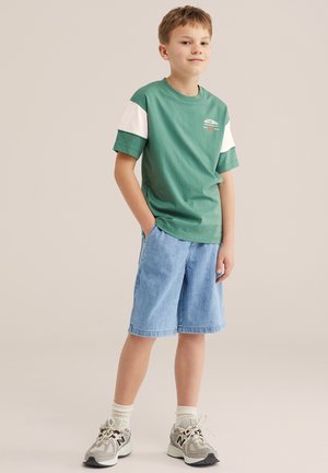 Poiss seisab käed taskutes, tal on seljas rohevalge lühikese varrukaga särk, helesinised teksapüksid, valged sokid ja hallid tossud.