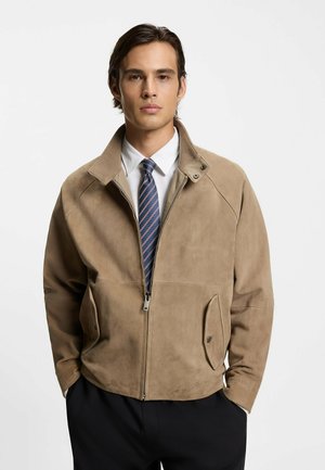 Giacca in camoscio color fango con collo alto, cerniera frontale e due tasche laterali. Abbinata a una camicia bianca e una cravatta a righe. Texture liscia.