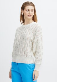 Strickpullover in Creme mit einem dekorativen Lochmuster, gerippten Bündchen und Saum, kombiniert mit blauen Hosen.