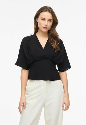 Kvinde iført en sort V-halsbluse med puffede korte ærmer og hvide højttaljede bukser, stående foran en ensfarvet baggrund.