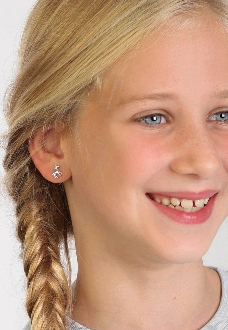 Zilveren studs in de vorm van een klein paard, met een gepolijste afwerking, gemonteerd op de oorlel van een kind met gevlochten haar.