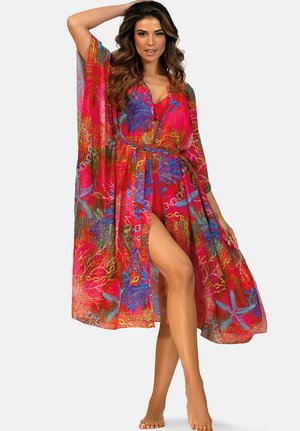 Kobieta o długich, falowanych włosach, ubrana w kolorową, zwiewną, czerwoną narzutkę plażową z niebieskimi wzorami rozgwiazd, pozująca boso na jednolitym tle.