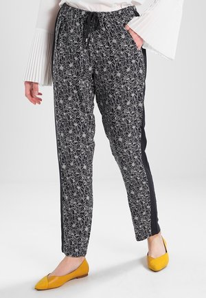 Pantalones negros con un patrón de contorno floral blanco y franjas laterales negras, combinados con zapatos planos amarillos y una blusa blanca con mangas acampanadas plisadas.