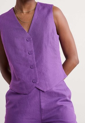 Personne portant un gilet violet sans manches boutonné et un pantalon violet assorti, les mains dans le dos.