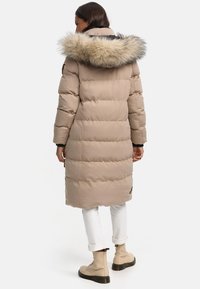 Beige Daunenjacke mit einer Kapuze aus Kunstfell, gesteppter Textur, knielangem Design und schwarzen Akzenten. Getragen mit weißen Hosen und beigen Stiefeln.