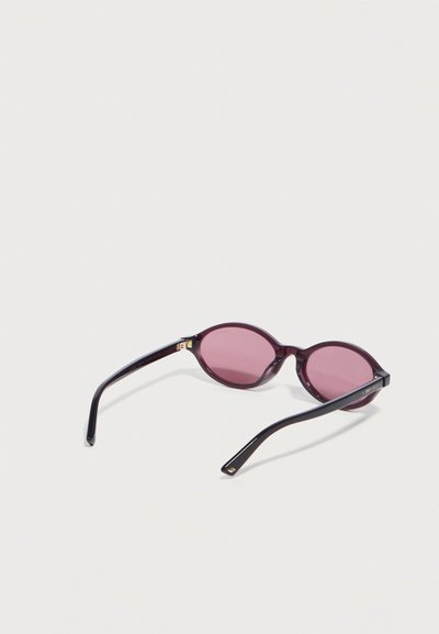Μαύρα οβάλ γυαλιά ηλίου με λεπτά πλαίσια και ροζ φακούς που ακουμπούν σε λευκή επιφάνεια.