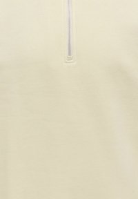 Hellbeiger Pullover mit einem Viertel-Reißverschluss-Kragen, hergestellt aus weichem Stoff. Er zeichnet sich durch eine glatte Textur und dezente Nähte rund um den Reißverschluss aus.