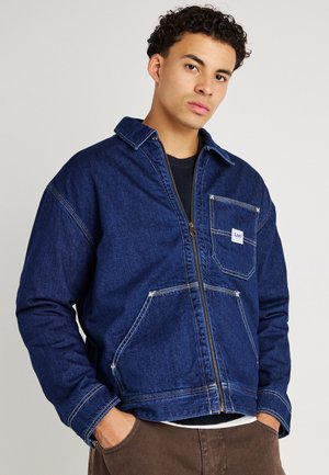 Chaqueta de mezclilla en azul oscuro con cierre de cremallera, cuello puntiagudo, dos bolsillos frontales y detalles de costura blanca en contraste.