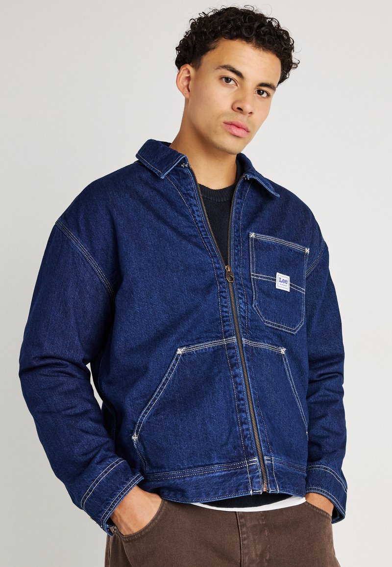 Denimjack in donkerblauw met een ritssluiting, spitse kraag, twee voorzakken en contrasterende witte stiksels.