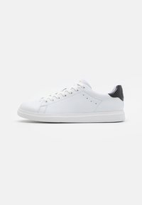 Sneaker en cuir blanc avec une finition lisse, bout rond, design à lacets, et un accent de talon noir. Présente des détails de coutures et des perforations.