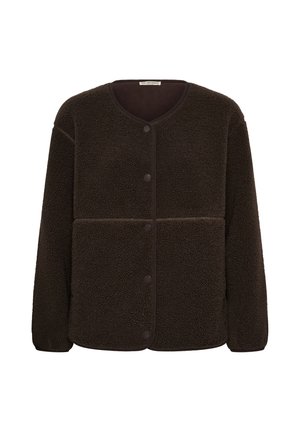 Femme souriante touchant ses cheveux, portant une veste polaire marron foncé boutonnée et un pantalon noir.