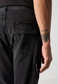 I pantaloni cargo neri in denim presentano tasche posteriori, una texture liscia e cuciture a vista. La cintura include una toppa in cuoio con l'etichetta "ANTONY MORATO."