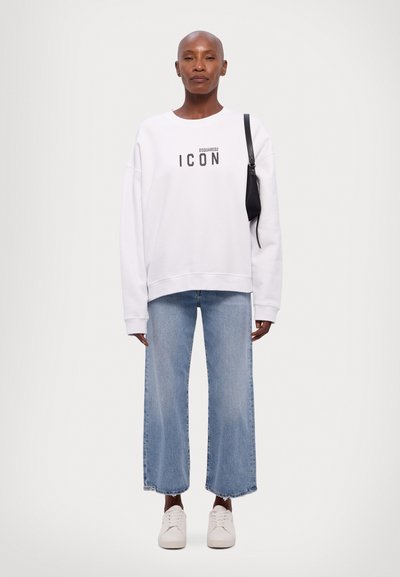 Sweatshirt oversized blanc avec un graphique "ICON", associé à un jean large bleu clair et des baskets blanches. Sac noir porté à l'épaule.
