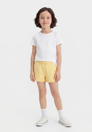Dieťa s tmavými kučeravými vlasmi, oblečené v bielom tričku, žltých vzorovaných šortkách, bielych ponožkách a bielych teniskách, stojace na bielom pozadí.