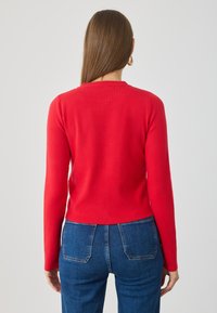 Roter Pullover mit langen Ärmeln aus glattem Strickstoff, mit rundem Ausschnitt und verkürztem Saum, kombiniert mit blauen Jeans.