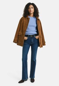 Veste en daim marron sur un pull en tricot bleu, associée à un jean évasé bleu foncé et des bottines marron. Fond blanc uni.