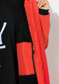 Veste rouge avec une doublure en polaire noire, présentant une poche zippée et des textures contrastées. Une main tient la veste ouverte pour révéler les détails.