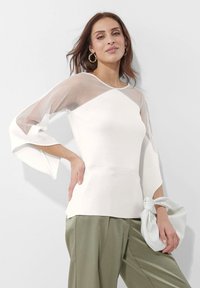 Witte langwaardige top met doorschijnende gaasaccenten, een getailleerde lijfstijl, flared mouwen en een gladde textuur. Gecombineerd met olijfgroene broeken en een witte tas.