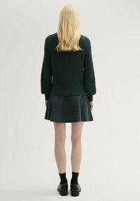 Suéter verde de canalé con mangas largas y falda acampanada de un oscuro color verde azulado. Combinado con calcetines negros y zapatos gruesos. Detalle de punto texturizado en el suéter.