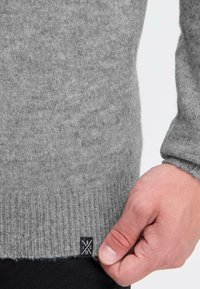 Szary sweter robiony na drutach z miękkiej wełny, z prążkowanym dołem i akcentującą metką z logo. Gładka tekstura z lekkim puszeniem.