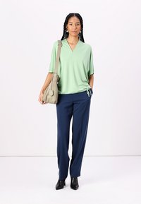 Top verde chiaro a maniche corte con scollatura a V e coulisse laterale, abbinato a pantaloni navy e stivali neri alla caviglia. Borsa a tracolla in tonalità neutra.