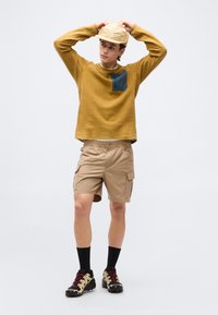 Νέος άντρας που ρυθμίζει το μπεζ καπέλο του, φοράει μουσταρδί κίτρινο πλεκτό πουλόβερ με μπλε τσέπη, χακί cargo σορτς, μαύρες κάλτσες και παπούτσια πεζοπορίας.