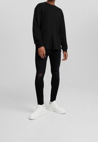 Hombre vestido con una camiseta de manga larga negra, jeans ajustados negros desgastados y zapatillas blancas, de pie contra un fondo liso.