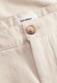 Svijetlo bež, strukturirana tkanina s metalnim kopčanjem; sadrži oznaku "GABBA" na vrhu, ističući svoj ležeran dizajn.