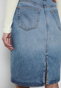 Jupe en denim avec un délavage bleu moyen, dotée de deux poches arrière, d'une fente au dos et d'une couture contrastée. Texture lisse avec une silhouette droite.