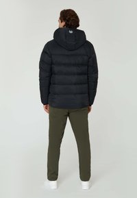 Sort pufferjakke med hætte, quilted tekstur og rundt logo på ryggen. Båret med olivengrønne bukser og hvide sneakers.