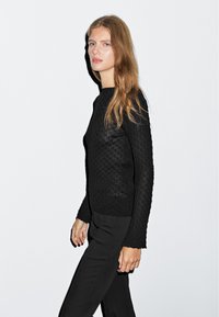 Black long-sleeve top with textured, quilted pattern; fitted shape, crew neckline, and ribbed cuffs. Paired with sleek black trousers.