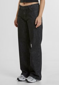 Jeans de mezclilla negra de pierna ancha con una cintura alta, costuras en contraste, diseño clásico de cinco bolsillos y cierre de botón. La textura parece lisa.