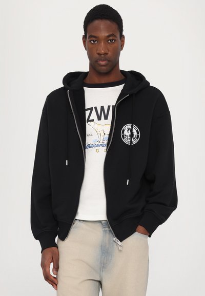 Black zip-up hoodie with a lined hood and front pockets, featuring a circular logo on the chest. Worn over a graphic t-shirt.