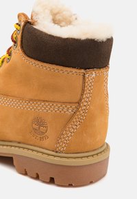 Botină din piele maro cu guler din blană sintetică crem, accente din suede maro, șireturi galbene și roșii, și o talpă din cauciuc texturat cu un logo de copac.