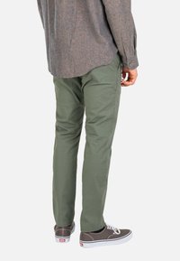 Person wearing green pants, gray button-up shirt, and gray low-top sneakers with white soles, standing against a white background.