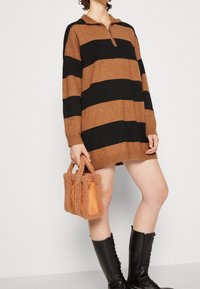 Maglione oversized a righe marroni e nere con colletto a metà zip, abbinato a una borsa marrone texturizzata con dettagli in pelle. Stivaletti neri alti fino al ginocchio.