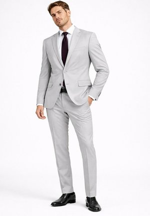 Homme debout portant un costume gris clair, une chemise blanche, une cravate sombre, une ceinture noire et des chaussures habillées noires sur fond blanc uni.