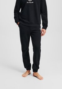 Hanorac negru cu manșete și tiv cu nervuri, având un logo din text alb. Pantaloni de trening negri asortati, cu manșete elastice și detaliu cu logo pe picior.
