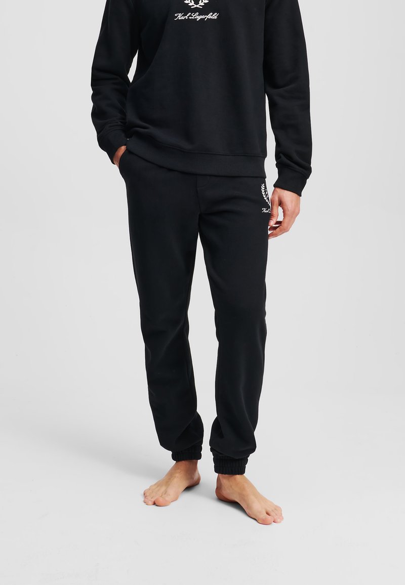 Hanorac negru cu manșete și tiv cu nervuri, având un logo din text alb. Pantaloni de trening negri asortati, cu manșete elastice și detaliu cu logo pe picior.