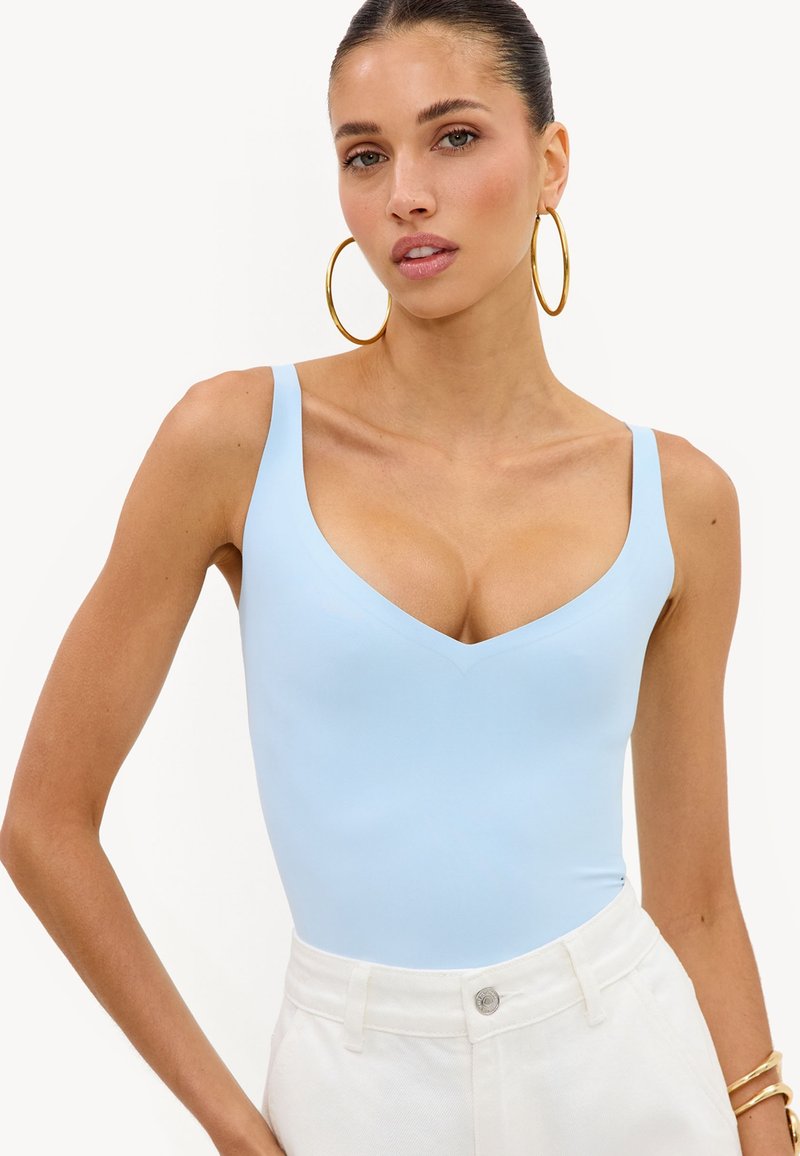 Femme aux cheveux attachés porte de grandes boucles d’oreilles dorées en anneau, un haut sans manches bleu clair et un pantalon blanc taille haute, posant devant un fond uni.