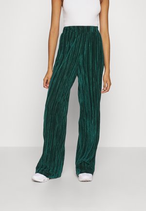 Vrouw draagt donkergroene fluwelen broek met hoge taille en wijde pijpen, gecombineerd met een wit mouwloos geribd topje en witte sneakers.