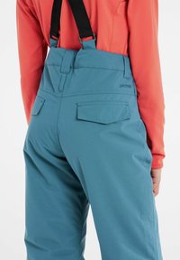 Blå skidbyxor med justerbara svarta hängslen, utrustade med två bakfickor och en slät yta, tillsammans med en klar orange topp.