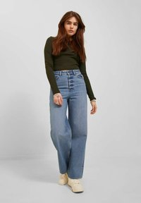 Farmerke od svijetloplave traper tkanine s visokim strukom i širokim krojem, s blagim izbljeđenjem, u kombinaciji s uskim dugim rukavima tamnozelene boje.