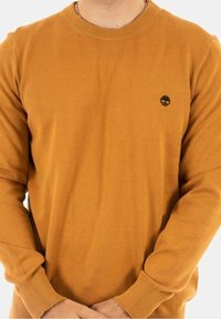 Pull en coton jaune moutarde avec un col rond, des poignets côtelés et un petit logo noir en forme d'arbre sur la poitrine. Texture lisse et coupe ajustée.