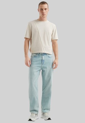 Beige T-Shirt mit kurzen Ärmeln und diagonalen Nahtdetails, kombiniert mit hellblauen Jeans im geraden Schnitt. Das Model trägt weiße und graue Sneaker.