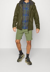 Veste imperméable vert olive, chemise à carreaux bleu et vert, shorts vert olive, chaussettes noires et chaussures de randonnée multicolores.